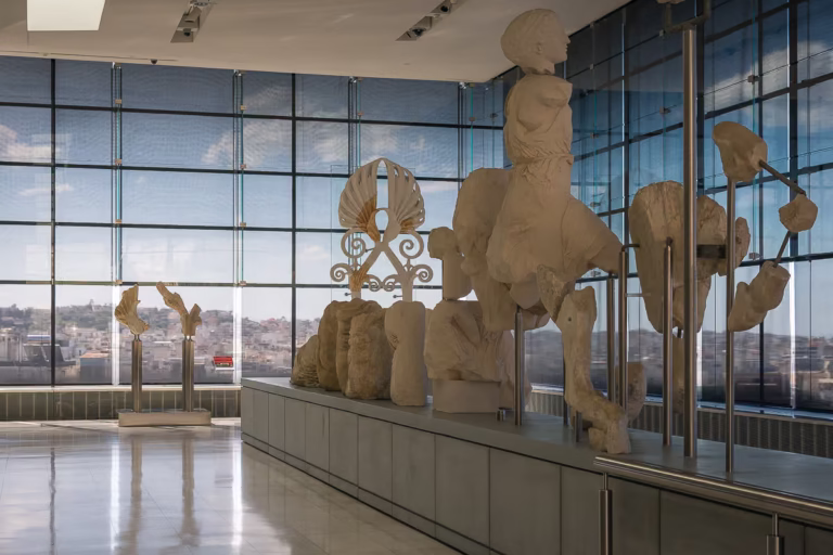 This image shows sculptures in a hall with large glass panels through which the view of Athens is visible.